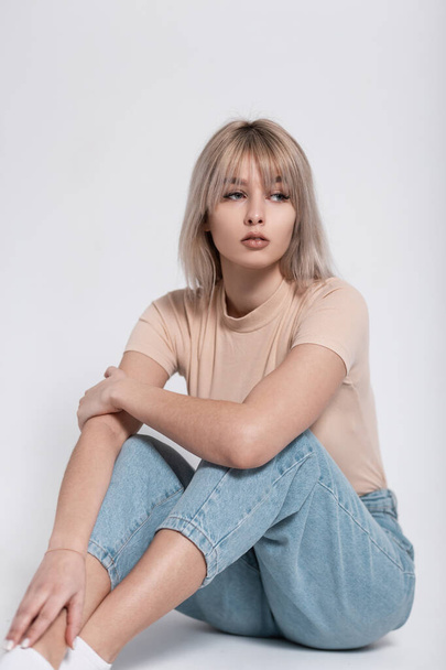 Cute trendy young woman fashion model with blond hair in a stylish T-shirt in fashionable blue jeans is resting near a white wall in the studio. Pretty beautiful girl. Modern youth wear for women. - Photo, Image