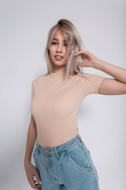 Mooie jonge vrouw in een stijlvol T-shirt in blauwe modieuze jeans met een trendy kapsel poserend bij een witte muur in de studio. Glamoureuze moderne mode model meisje binnen. Jongerenlente stijl. - Foto, afbeelding