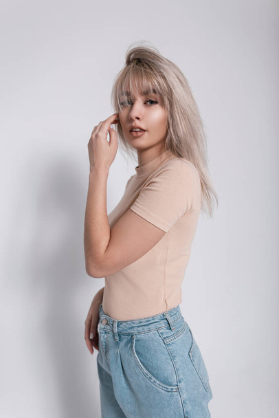 Fashion model of a glamorous young blonde woman in stylish blue jeans in a trendy shirt with a hairstyle stands near a white wall in the room. Modern beautiful elegant girl in youth fashionable wear.  - Φωτογραφία, εικόνα