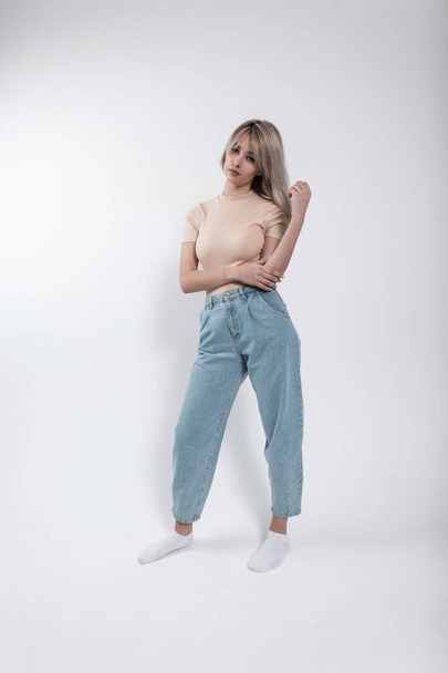 Beautiful young blonde woman in a fashionable beige shirt in vintage blue jeans stands near a modern white wall indoors. Pretty nice girl fashion model posing in the studio. Casual womens wear. - 写真・画像
