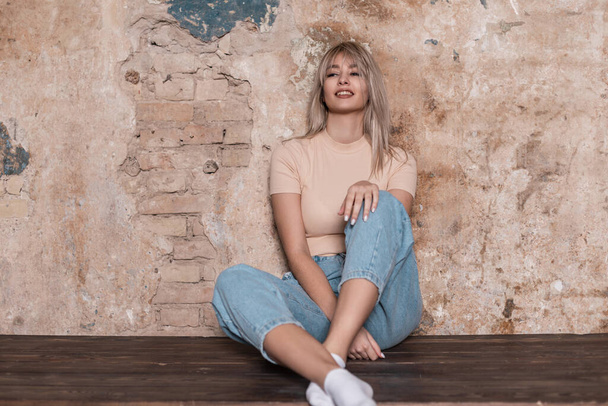 Young trendy model of a positive beautiful woman with a charming smile in fashionable clothing is resting sitting near a vintage brick wall in the room. Attractive happy blonde girl posing indoors. - Photo, Image