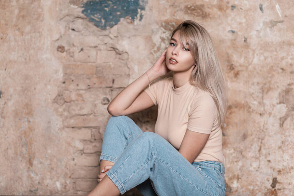 Modern pretty young woman sits near a vintage brick wall in the studio. Beautiful pretty fashionable girl model with sexy lips in stylish clothes rests indoors. Trendy European lady in youth wear. - 写真・画像