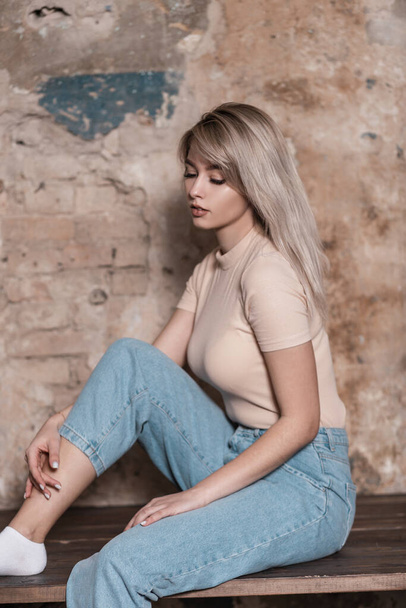 Pretty fashion young blonde woman in stylish blue jeans in a trendy shirt poses near a brick old wall indoors. Glamorous girl model in casual clothes sits and looks down. Beauty lady in the studio. - Photo, Image