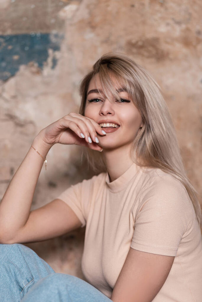 Portrait of a fashionable happy young woman with positive beautiful smile in a vintage T-shirt in blue trendy jeans on a background brick wall in the room. Pretty cheerful girl blonde sits indoors. - Foto, afbeelding