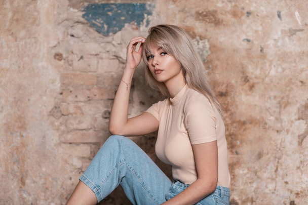 Europese mooie jonge blonde vrouw blond met sexy lippen in een modieus beige t-shirt met een stijlvolle jeans zit in de buurt van een bakstenen muur in de kamer. Aantrekkelijk trendy meisje mode model rust binnen.  - Foto, afbeelding