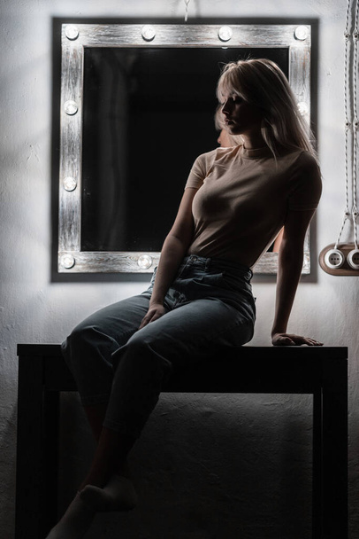 Fashionable pretty young woman model in a dark room. Sexy elegant blonde girl in stylish clothes sits on a table near a vintage white mirror for makeup with bright lamps in the evening in the bedroom. - Фото, изображение