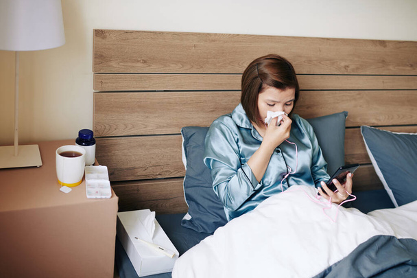 Kranke junge Frau bleibt zu Hause, liegt im Bett, pustet sich die Nase und checkt soziale Medien auf dem Smartphone - Foto, Bild