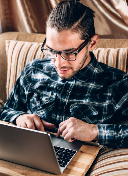 Een knappe jonge freelancer in een blauw shirt en bril met diopters werkt thuis op een zilveren laptop. Afstandswerk tijdens de quarantaine. - Foto, afbeelding