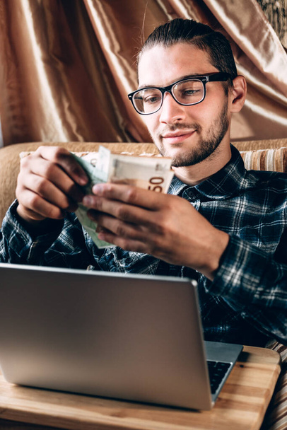 A handsome young man in a blue shirt and glasses works at a silver computer and holds Euro paper money in his hands. Remote work for a freelancer in a comfortable home environment, online shopping. - Foto, afbeelding