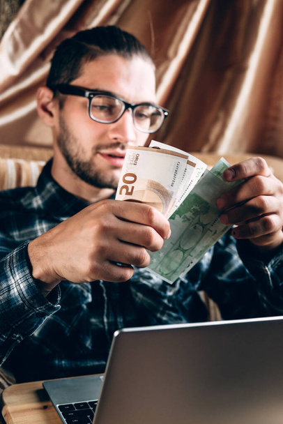 A handsome young man in a blue shirt and glasses works at a silver computer and holds Euro paper money in his hands. Remote work for a freelancer in a comfortable home environment, online shopping. - Foto, immagini
