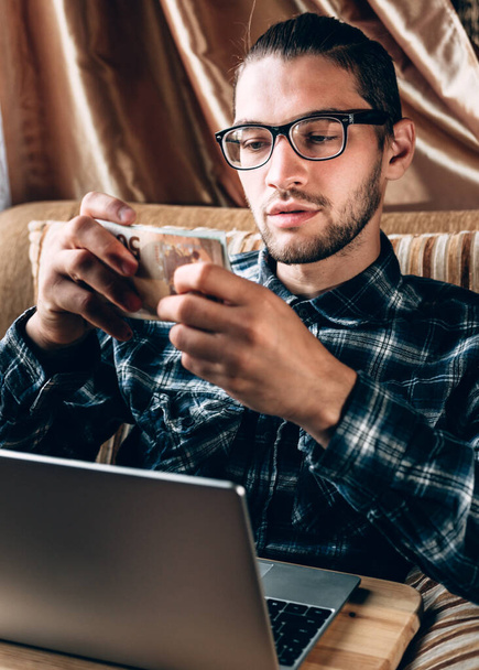 Hezký mladý muž v modré košili a brýlích pracuje u stříbrného počítače a v rukou drží europapírové peníze. Vzdálená práce na volné noze v příjemném domácím prostředí, online nakupování. - Fotografie, Obrázek