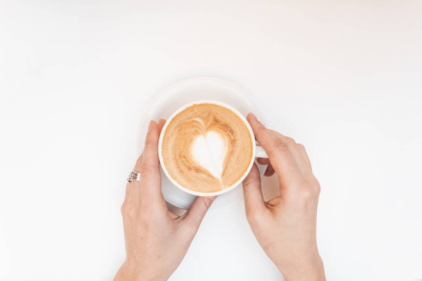 Closeup of female hands with a vintage cup of cappuccino with cute pattern on white background. Morning coffee break in a coffee shop. View from above. - Photo, Image