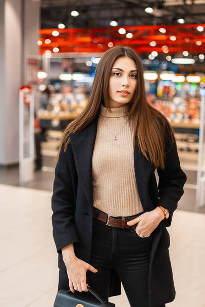 European young modern woman brunette with long hair in trendy black coat with fashionable leather bag posing in modern shopping mall. European girl walks on the shop. Pretty urban lady fashion model. - Fotó, kép