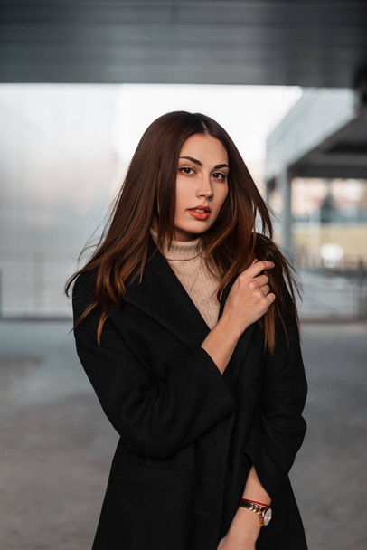 Portrait young attractive woman with luxurious long brown hair with sexy lips with natural makeup in an elegant black coat near a luminous modern building. Cute girl fashion model on the street. - Foto, immagini