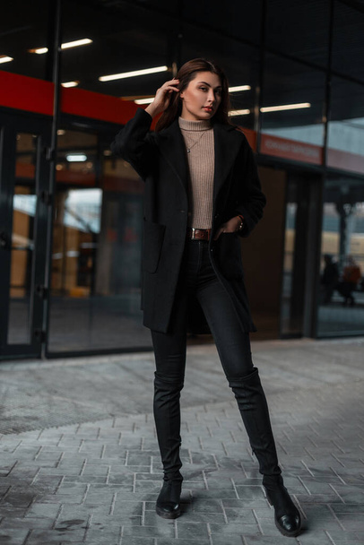 Hübsche junge brünette Frau in modischem schwarzen Mantel, beigem Hemd, Jeans und Schuhen steht neben einem modernen Glasgebäude im Freien. Schlanke trendige Mädchen-Mode-Modell in stilvoller Kleidung auf der Straße. - Foto, Bild