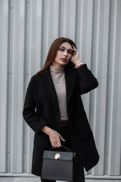 Elegant urban model young woman with long hair in shirt in luxurious fashionable black coat with leather handbag posing near metal silver wall on the street. Beautiful girl model. Cute trendy lady. - Photo, Image
