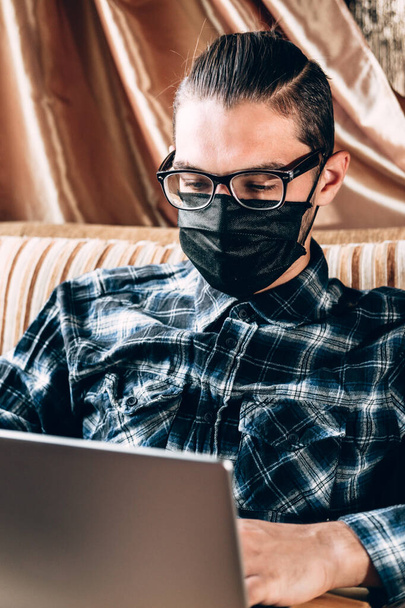 Ein gutaussehender junger Freiberufler mit Dioptrienbrille und schwarzer Schutzmaske arbeitet zu Hause auf einem silbernen Laptop. Fernbedienung während der Quarantäne, um den Text auszudrucken und wichtige Geschäfte zu tätigen. - Foto, Bild
