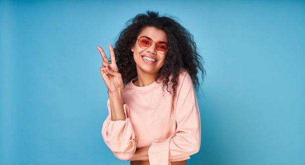 The attractive, cheerful young black girl, with long black curly hair, shows a pacifist sign, wearing a kejual blouse and red sleepwalking glasses. Copy space. - Foto, Bild