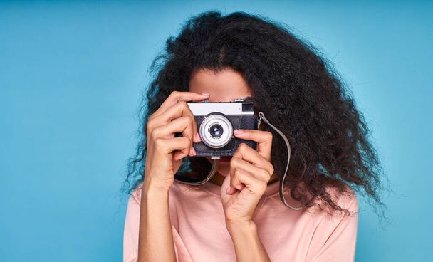 A close-up, curly brunette in a beige blouse, takes a picture on a retro camera, posing in the studio on a blue background. - Фото, изображение