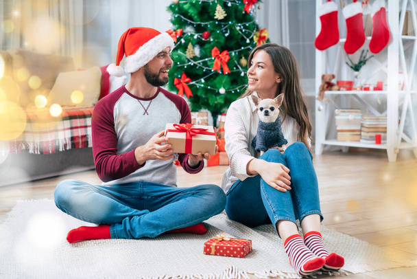 Een romantisch kerstcadeau. Gelukkig mooi jong paar in liefde zijn het maken van een verrassing en het geven van geschenken voor elkaar, terwijl zitten op de vloer thuis op kerstboom achtergrond. - Foto, afbeelding