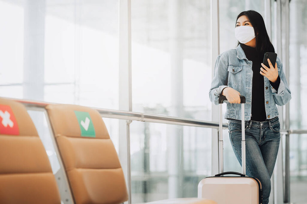 Gelukkig aziatische vrouw reiziger dragen masker ter bescherming tegen coronavirus houden smartphone staande met bagage naast sociale afstandelijke stoel - Foto, afbeelding