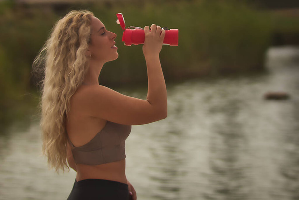 Young blonde woman drinking water after running from a pink plastic bottle with background water in a park. - Photo, Image