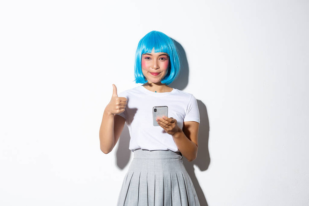 Portrait of satisfied smiling pretty girl in halloween costume, showing thumbs-up in approval of something in smartphone, standing over white background - Photo, Image