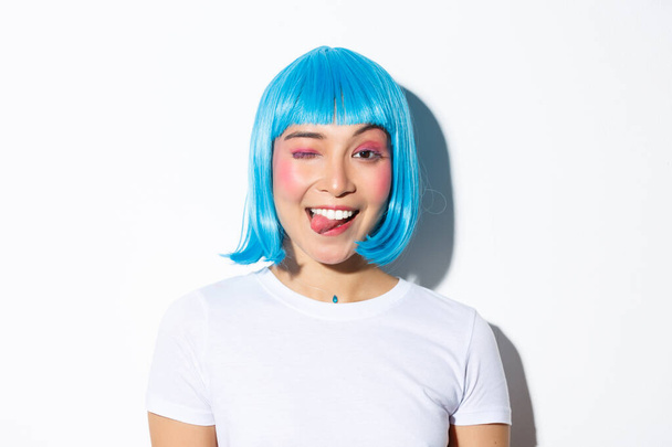 Крупный план привлекательной тусовщицы в голубом парике, показывающей язык и подмигивающей счастливой, празднующей Хэллоуин, стоящей на белом фоне - Фото, изображение