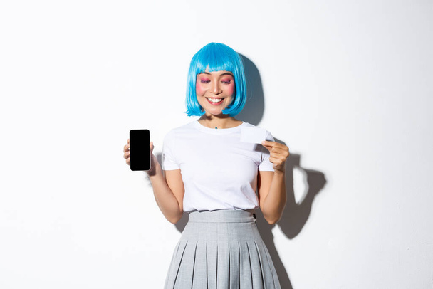 Kaunis hymyilevä aasialainen tyttö sinisessä peruukissa näyttää tyytyväiseltä luottokortilla, näyttää älypuhelimen näytön, seisoo valkoisen taustan yläpuolella - Valokuva, kuva