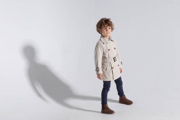 Full length of a stylish adorable curly little boy, wear in a trench, poses in studio, isolated white background. - Foto, imagen