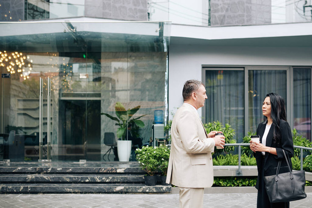 Серйозні бізнесмени, що стоять за межами офісної будівлі, розмовляють і п'ють, виймають каву під час перерви
 - Фото, зображення