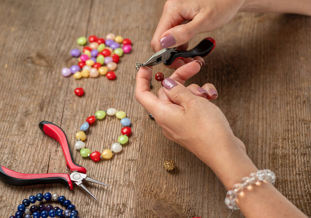 Jewelry making. Making a bracelet of colorful beads. Female hands with a tool on a rough wooden table. Selective focus - Photo, Image