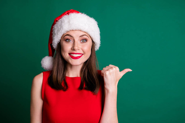 Clsoeup photo of attractive charming crazy lady good mood excited direct thumb finger side empty space shopper wear red snow girl dress fluffy santa cap isolated green color background - Zdjęcie, obraz
