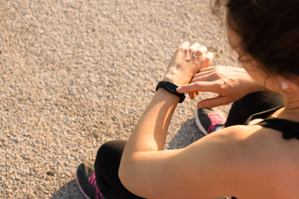 Athlète femme touchant suivi de forme physique vérifier le pouls ou des informations de sport, réglage de la montre intelligente avant de courir ou séance d'entraînement du matin, assis à l'extérieur dans la journée ensoleillée d'été. - Photo, image