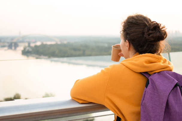 Back view of young brunette woman tourist in yellow hoodie with pink backpack drinking coffee to go and enjoying city and river view standing on bridge during the walk. Travel concept. Copy space - Photo, Image