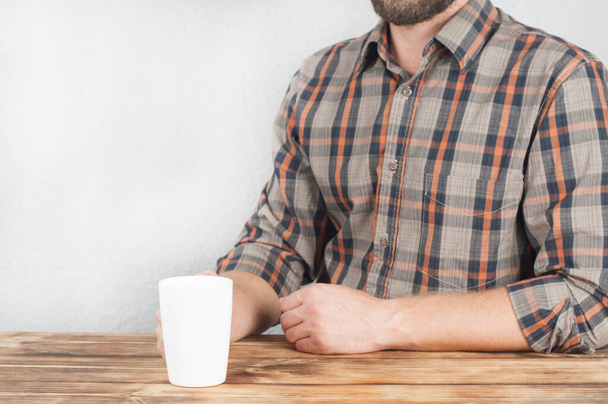 A young man in a shirt holds a cup in his hand. Office worker. Sitting at a wooden table - Фото, изображение