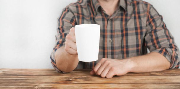 A young man in a shirt holds a cup in his hand. Office worker. Sitting at a wooden table - Foto, Imagen