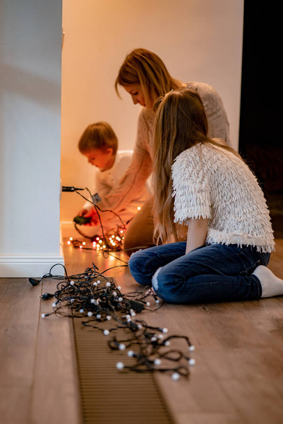 Maman et les enfants décorent le sapin de Noël avec des guirlandes et des jouets. La famille décore le sapin de Noël. Se préparer pour Noël et le Nouvel An. Les enfants se réjouissent devant l'arbre. - Photo, image