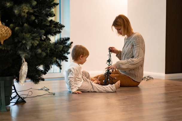 Mom and children decorate the Christmas tree with garlands and toys. The family decorates the Christmas tree. Preparing for Christmas and New Years. Children rejoice at the tree. - Photo, image