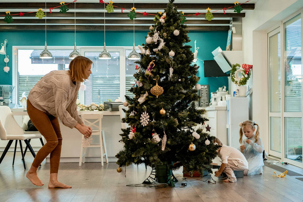 Mom and children decorate the Christmas tree with garlands and toys. The family decorates the Christmas tree. Preparing for Christmas and New Years. Children rejoice at the tree. - Φωτογραφία, εικόνα