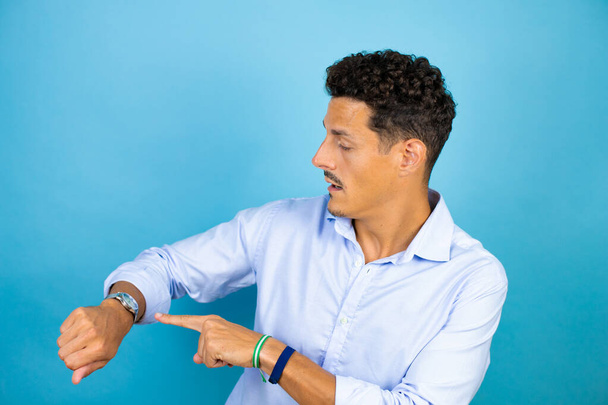 Young handsome man wearing blue shirt over isolated blue background surprised and pointing his watch because it's late - Photo, Image