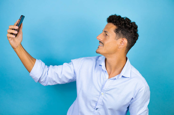 Молодой красивый мужчина в голубой рубашке на изолированном синем фоне делает селфи с телефоном - Фото, изображение