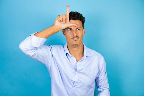 Jonge knappe man draagt blauw shirt over geïsoleerde blauwe achtergrond lachen van mensen met vingers op het voorhoofd doen verliezer gebaar bespotten en beledigend. - Foto, afbeelding