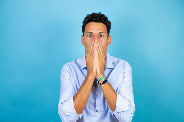 Junger gutaussehender Mann trägt blaues Hemd über isoliertem blauem Hintergrund, die Hände über dem Mund und überrascht - Foto, Bild