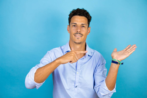 Jeune homme beau portant chemise bleue sur fond bleu isolé étonné de regarder la caméra présentant quelque chose sur la main et pointant du doigt. - Photo, image