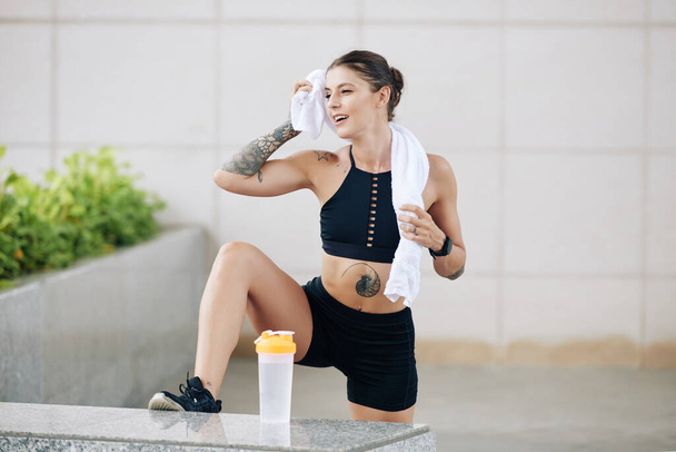 Glimlachende fitte jonge vrouw in top en korte broek vegen af zweet na intensieve training - Foto, afbeelding