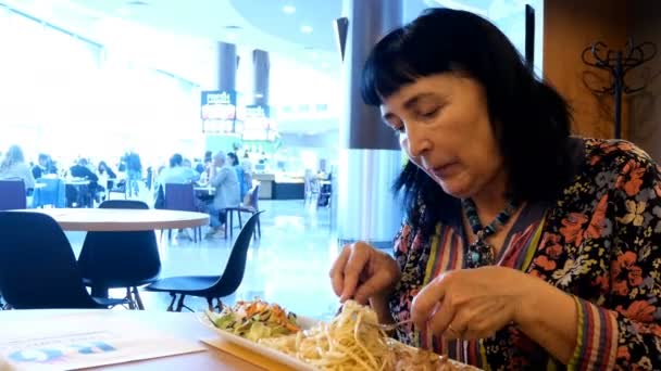 Kypsä aikuinen nainen syö pastaa salaatilla ja lihalla kahvilassa tai ravintolassa ostoskeskuksessa haarukalla ja veitsellä. lähikuva. - Materiaali, video