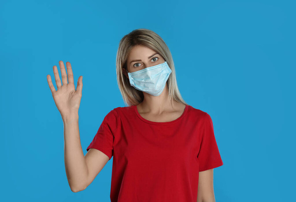 Woman in protective mask showing hello gesture on light blue background. Keeping social distance during coronavirus pandemic - Foto, Bild