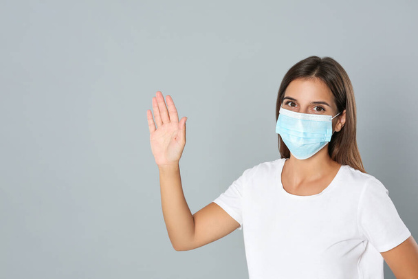 Woman in protective mask showing hello gesture on grey background, space for text. Keeping social distance during coronavirus pandemic - Photo, image