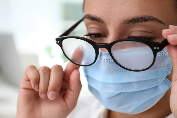 Woman wiping foggy glasses caused by wearing medical mask indoors, closeup - Фото, изображение